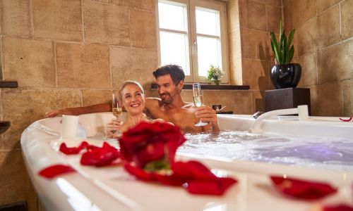 Pärchen sitzt zusammen in Badewanne, welche mit roten Rosenblättern dekoriert ist. Beide haben ein Glas Sekt in der Hand und lächeln