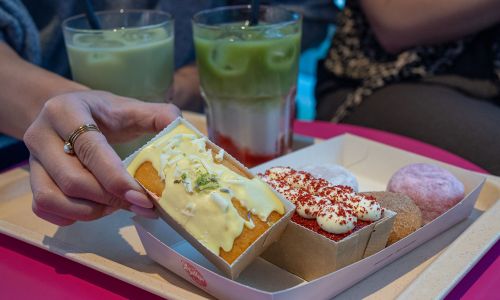 Nahaufnahme von einem Tablett mit drei Mini-Donuts, zwei kleinen Küchlein und zwei Iced Strawberry Matcha Latte in Sallys Café im Isländischen Bereich des Europa-Park. Eine Hand hält eins der kleinen Küchlein.